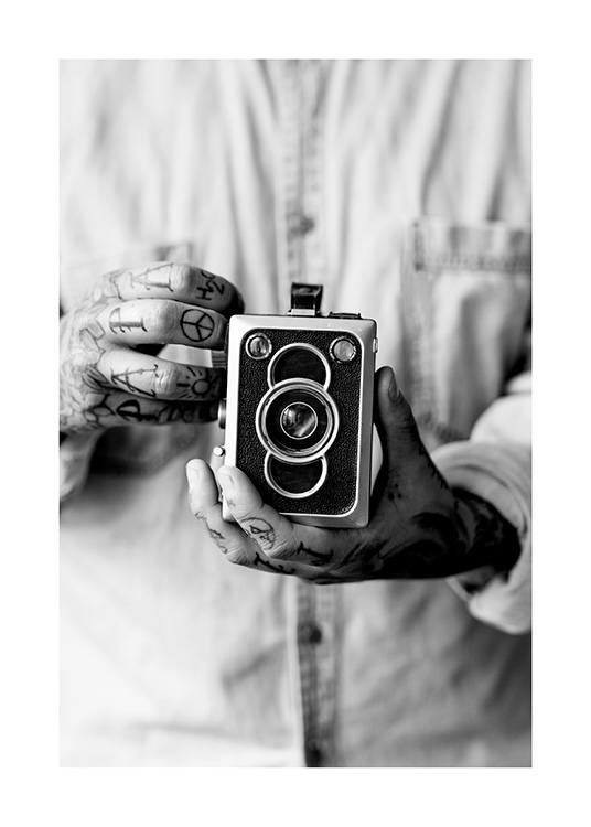  – Black and white photograph of a vintage camera being held by a man with tattoed hands