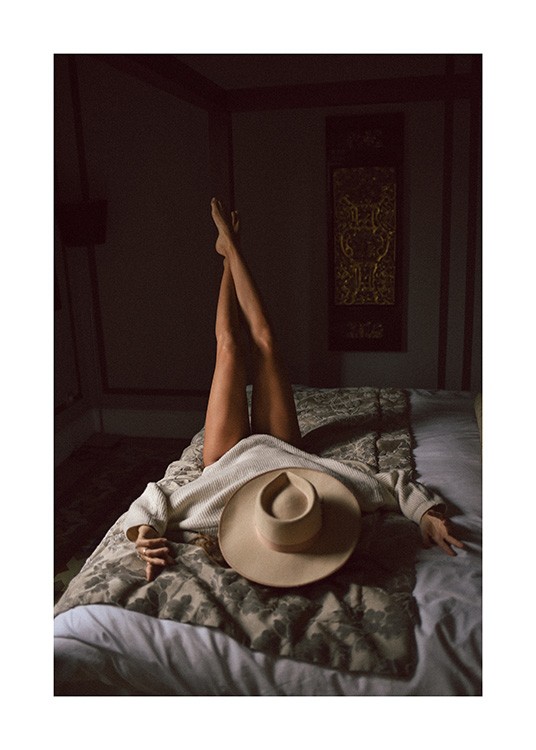  – Photograph of a woman laying on a bed with her legs stretched up into the air and a hat covering her face