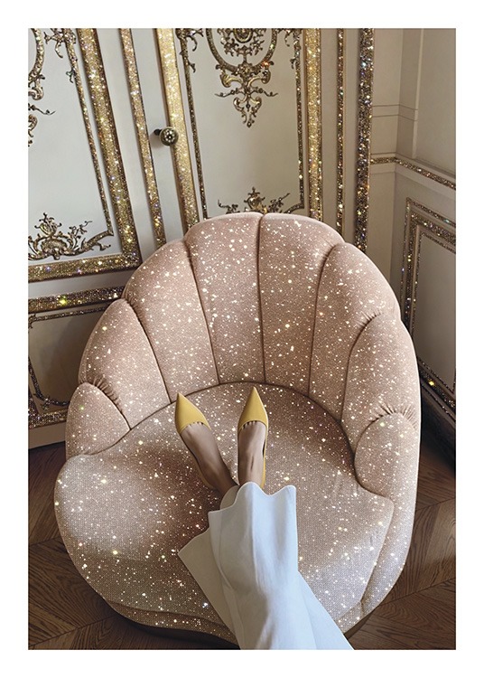  – Photograph of a pink, sparkling chair in front of a gold and white wall, with a woman resting her feet on the chair