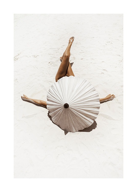  – Photograph of a woman covered by a beige parasol, laying on white sand with her legs crossed