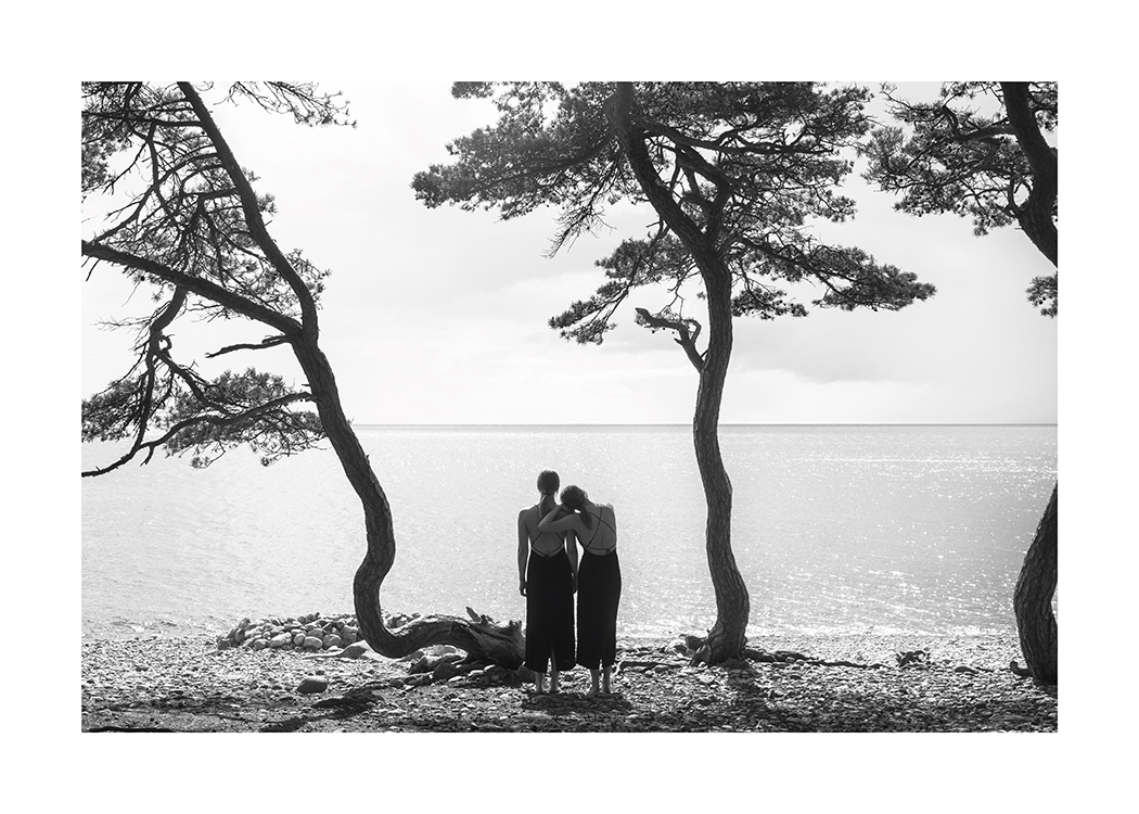  – Black and white photograph of a pair of women looking out at the water from a beach with trees